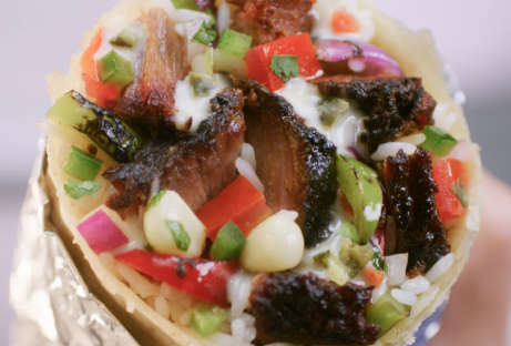 A close up point of view photo of a Smoked Brisket Burrito wrapped in foil and filled with white rice, fajita veggies, hand-chopped Smoked Brisket, fresh tomato salsa, roasted chili corn salsa, and sour cream.